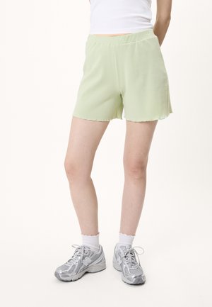 Person iført lysegrønne ribstrikkede shorts, hvide sokker, sølvfarvede sneakers og en hvid ærmeløs top mod en ensfarvet baggrund.