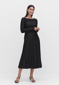 Mustapaitainen mekko, jossa on pitkät hihat ja kellotettu helma, ja joka on varustettu tekstuuripinnalla. Yhdistetty mustiin nilkkaremmikenkiin.