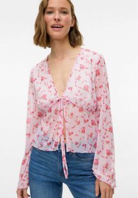 Blouse florale en tissu léger et transparent rose, avec des manches longues, un décolleté en V profond et un accent à nouer à l'avant. Présente de petites fleurs multicolores.