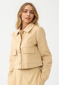 Beige verkürzte Jacke mit spitzem Kragen, großen Fronttaschen und Knopfverschluss, aus einem glatten, gewebten Stoff gefertigt.