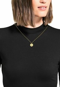 Gul cirkulär hängsmycke på en tunn guldkedja, bärs över en svart hög nacktop, med en texturerad yta och intrikat design.