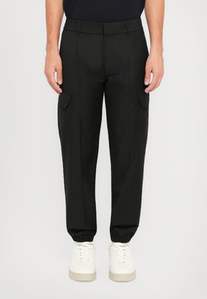 Sorte cargo bukser med elastisk linning, avsmalnende mansjetter og sidelommer. Laget av et glatt, lett stoff. Brukt med hvite sneakers.