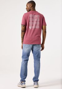 Camiseta de manga corta color burdeos con un patrón gráfico gris en la parte posterior. Combinada con jeans de mezclilla azul claro y zapatillas grises con acentos azules.