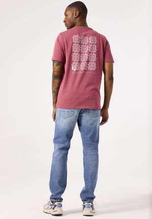 Korte mouw, bordeauxrode t-shirt met een grijs grafisch patroon op de achterkant. Gecombineerd met lichtblauwe spijkerbroek en grijze sneakers met blauwe accenten.
