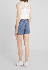 Top bianco senza maniche con dettagli in pizzo sul retro, abbinato a pantaloni corti blu a fantasia. La modella indossa sneakers bianche alte. Outfit casual.