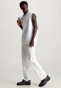 White sleeveless sweatshirt with embossed logo, paired with loose-fitting white pants and black shoes. Smooth fabric and clean design.