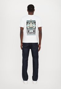 Camiseta blanca con diseño gráfico que presenta una cara, acentos azules y texto en la parte posterior. Combinada con jeans de mezclilla oscura y zapatillas blancas.