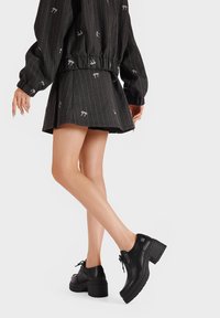 Scarpe in pelle nera con tacco spesso, punta arrotondata e lacci, abbinate a una gonna nera a righe sottili con piccoli motivi a fiocco bianchi.