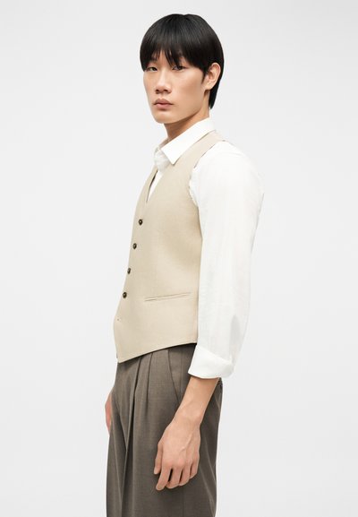 Νέος άνδρας με ίσια μαύρα μαλλιά, που φοράει μπεζ γιλέκο πάνω από λευκό πουκάμισο και καφέ πλισέ παντελόνι, στέκεται μπροστά σε λευκό φόντο.