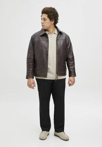 Brown leather jacket with a zip closure, light beige sweater underneath, black pants, and beige slip-on shoes. Model stands sideways.