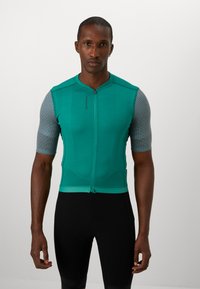 Maglia da ciclismo da uomo, color teal con tessuto texturizzato, maniche corte in mesh grigio, zip frontale e design aderente, abbinate a pantaloni corti da ciclismo neri.