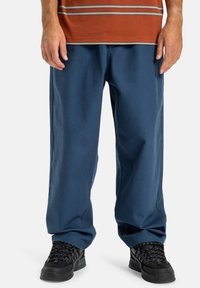 Modré bavlněné kalhoty s volným střihem, mírně zúžené nohavice, nošeny s černými texturovanými teniskami a hnědým pruhovaným tričkem.