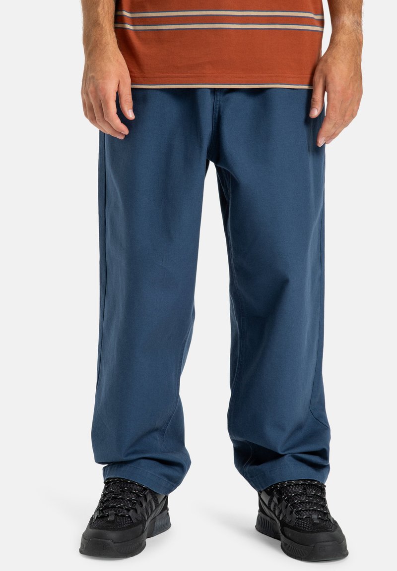 Modré bavlněné kalhoty s volným střihem, mírně zúžené nohavice, nošeny s černými texturovanými teniskami a hnědým pruhovaným tričkem.