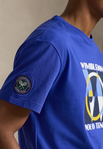 Άτομο που φοράει μπλε t-shirt τένις Polo Wimbledon με κηλίδα λογότυπου Wimbledon στο μανίκι και πολύχρωμο γραφικό στο στήθος.