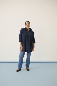 Femeie care stă pe un fundal simplu, purtând o cămașă bleumarin cu nasturi, cu un pulover asortat aruncat peste umeri, blugi albaștri și pantofi maro.