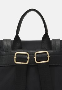 Sac à dos noir en nylon mat, avec une poignée supérieure arrondie, des bretelles ajustables ornées de boucles en métal doré, et des accents texturés.