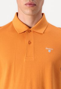 Πορτοκαλί πόλο μπλουζάκι με κλασικό γιακά, δύο κουμπιά και μικρό λογότυπο στο στήθος. Υφασμάτινη δομή με ομοιόμορφο χρώμα.