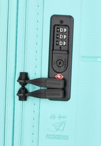 Angolo di una valigia turchese con lucchetto a combinazione; due cerniere con maniglie nere; superficie testurizzata; logo in rilievo e dettaglio del marchio di viaggio.