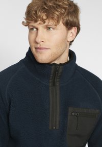 Navy fleece quarter-zip pullover with a black zippered chest pocket and contrast fabric, featuring a soft, textured surface.