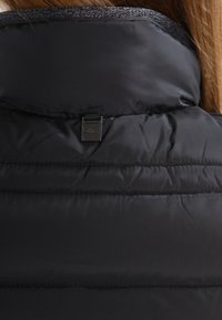 Czarna kurtka puchowa z wysokim kołnierzem, przeszytymi sekcjami dla lepszej izolacji oraz małą metką z logo z tyłu, blisko szyi.