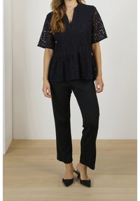 Top peplum in pizzo nero con maniche corte e scollatura a V. Abbinato a pantaloni neri su misura e tacchi a slitta neri, su un pavimento in legno chiaro.