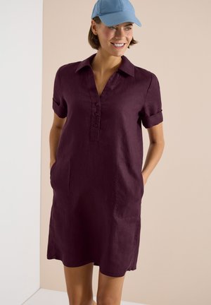 Femme souriante portant une robe chemise prune à manches courtes avec poches avant et une casquette de baseball bleu clair, debout devant un fond neutre.