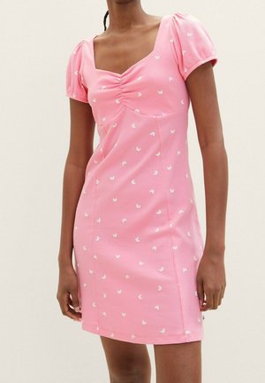 Kobieta ubrana w krótką różową sukienkę z bufkowatymi rękawami i marszczonym dekoltem, ozdobioną małymi białymi wzorami motyli.