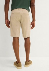 Pantaloni corti beige su misura con due tasche posteriori, abbinati a sneakers marroni. Il tessuto sembra leggero e liscio, adatto per il clima caldo.