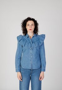 Camicia di denim con un ampio colletto arricciato, maniche lunghe e chiusura con bottoni. Indossata con jeans in denim abbinati, che mostrano una palette di blu coesa.