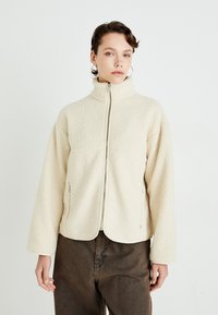 Young woman wearing cream zip-up fleece jacket and brown pants, standing against a plain white background, facing forward.