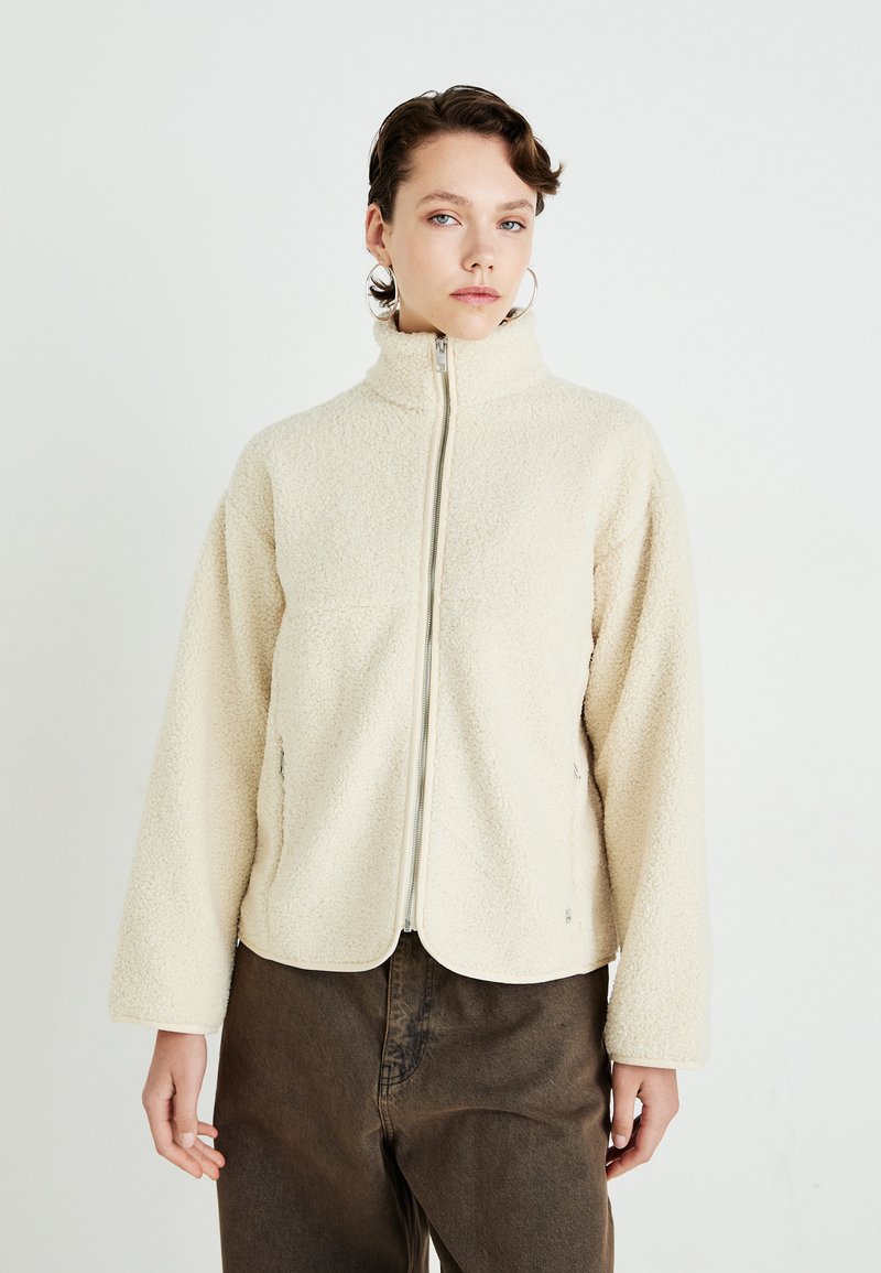 Young woman wearing cream zip-up fleece jacket and brown pants, standing against a plain white background, facing forward.