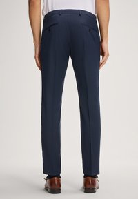 Marineblauwe op maat gemaakte broek met een gladde textuur, voorzien van twee achterzakken met knopen en een recht silhouet. Bruine leren schoenen zichtbaar.