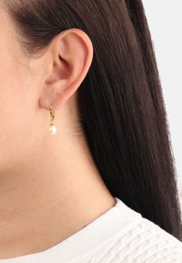 Guldpläterad hoopörhänge med en ensam vit pärla som hänger. Närbild på örat, som visar släta hår och en strukturerad vit klädnad.