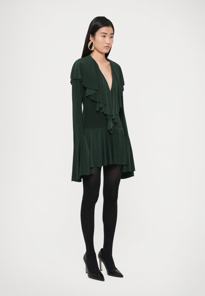 Zielona sukienka z falbankami, głęboki dekolt w serek, długie rękawy i rozkloszowany dół; w zestawieniu z czarnymi, kryjącymi rajstopami oraz czarnymi, spiczastymi szpilkami.