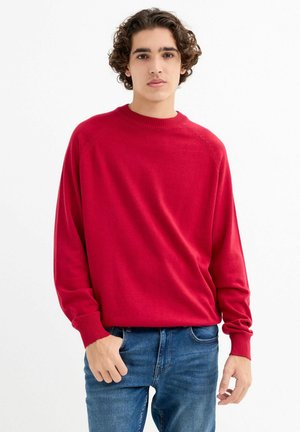 Jersey de punto rojo con cuello redondo, puños acanalados y un ajuste relajado. Combinado con jeans azules, presentado sobre un fondo blanco.