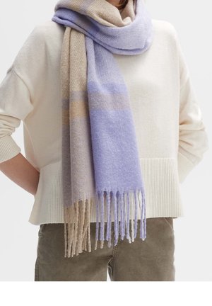 Persoon die een crèmekleurige trui en taupekleurige broek draagt, met een lange gevederde sjaal in beige en lavendel om de nek gewikkeld.