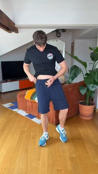 Mann in schwarzem T-Shirt und marineblauer Shorts stellt den Bund im Wohnzimmer mit Holzfußboden, orangem Sofa, Pflanzen und Fernseher ein.