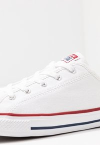 Białe płótno trampki z przodem na sznurowadła, z metalowymi oczkami, czerwono-niebieską paskowaną podeszwą i małym logo na języku.