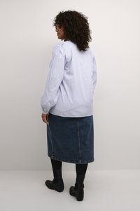 Blouse rayée bleu et blanc avec des manches bouffantes, associée à une jupe en denim foncé. Le mannequin porte des bottines noires. Fond neutre.
