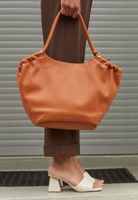 Borsa tote in ecopelle marrone con cima arricciata, dettagli dorati e texture morbida. Abbinata a sandali con tacco a blocco di colore chiaro e pantaloni marroni.