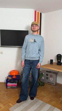Ljusblå sweatshirt med hundmotiv, lösa blå jeans, mörkbruna skor och en beige keps bak och fram. Står inomhus mot en vit vägg.