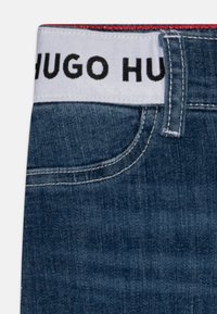 Modré džíny vyrobené z denimu s bílým pasem, na kterém je černý nápis "HUGO". Na pasu jsou výrazné červené švy.