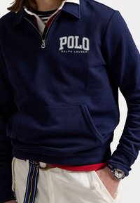 Námořnická modrá mikina s kapucí a zipem, bílý límeček a přední kapsa. Na přední straně je logo "POLO RALPH LAUREN" v bílé barvě. Materiál je směs bavlny.
