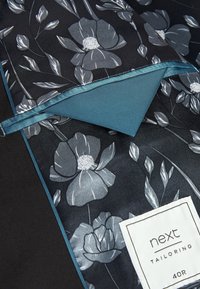 Sort stof med et blomstermønster i grå og hvid. Akvamarin satinaccenter og etiket med teksten "next tailoring 40R".