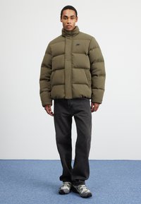 Giacca a vento verde oliva con colletto alto, caratterizzata da un motivo cucito, chiusura con zip e logo nero, abbinata a jeans neri e sneaker grigie.