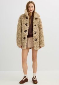 Joven mujer viste un abrigo de piel sintética rizado beige, falda color canela, sudadera marrón, calcetines blancos y zapatillas marrones, de pie contra un fondo blanco.