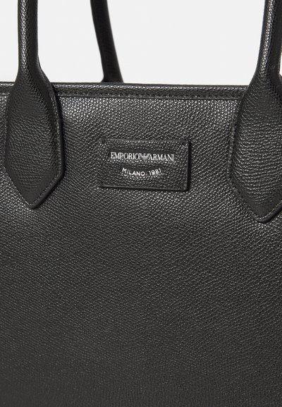 Svart läder tote-väska med en texturerad yta. Har en logotyp patch som står "EMPORIO ARMANI MILANO 1981." Har två robusta handtag.