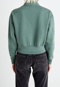 Felpa verde con orlo e polsini a costine, abbinata a jeans neri con tasche posteriori e etichetta del marchio visibile.