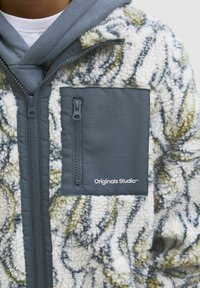 Giacca in pile con un motivo testurizzato bianco e verde, dotata di una tasca in tessuto grigio con zip e logo "Originals Studio".