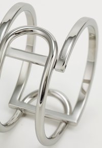Pulsera de brazalete de metal plateado con bucles entrelazados y formas geométricas. Presenta un acabado pulido y un logo grabado.